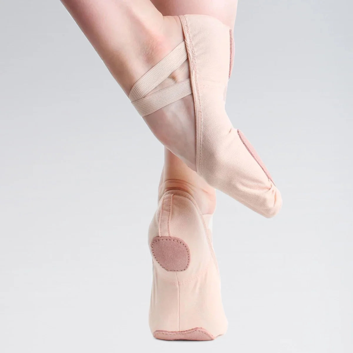 A close-up of a dancer's feet wearing pale blush pink ballet pointe shoes with elastic crisscross straps, shown against a light gray studio background.