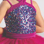Young dancer wearing a raspberry pink sequin tutu costume by 1st Position featuring a crossover halterneck design, sparkling sequin bodice and layered tutu skirt with blue glitter trim.
