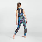 A woman modeling a sleeveless high-neck athletic jumpsuit from the back, featuring a black mesh back panel and a vibrant blue, purple, and green abstract swirl pattern with black mesh leg panels, set against a light gray studio background.