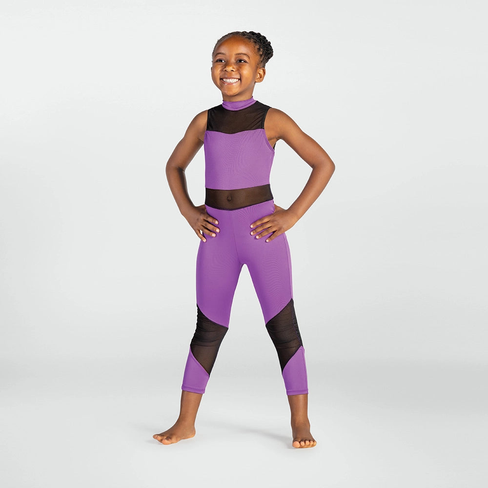 A young girl stands barefoot against a light gray studio background, wearing a purple and black athletic two-piece outfit consisting of a sleeveless crop top and matching leggings.