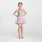 A young girl stands barefoot against a light gray studio background, wearing a sleeveless dance or skating dress with a pastel floral bodice, a light blue waistband, and a pale pink flared skirt with a light blue hem.