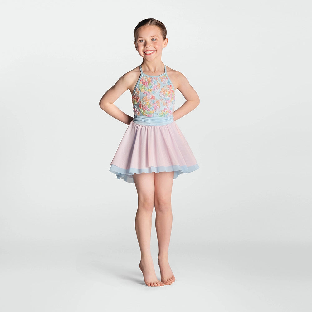 A young girl stands barefoot against a light gray studio background, wearing a sleeveless dance or skating dress with a pastel floral bodice, a light blue waistband, and a pale pink flared skirt with a light blue hem.