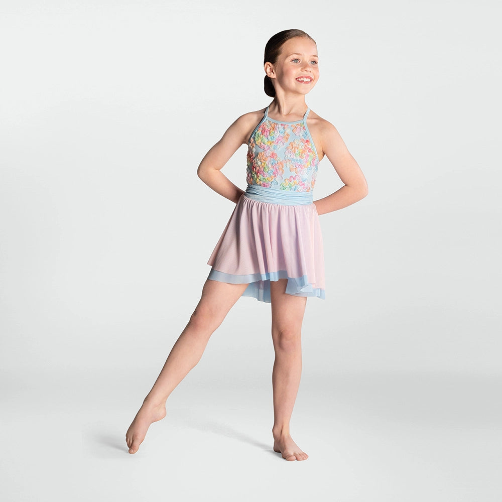 A young girl dancer posing in a sleeveless halter-neck leotard dress with a multicolor floral patterned bodice, a light blue waistband, and a layered pink and light blue mesh skirt, set against a plain light gray studio background.