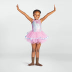A young girl with braided hair stands in a ballet pose with arms raised, wearing a sparkly lavender and silver sequined leotard with a pink tutu skirt trimmed in light blue, paired with brown dance socks, against a light gray studio background.