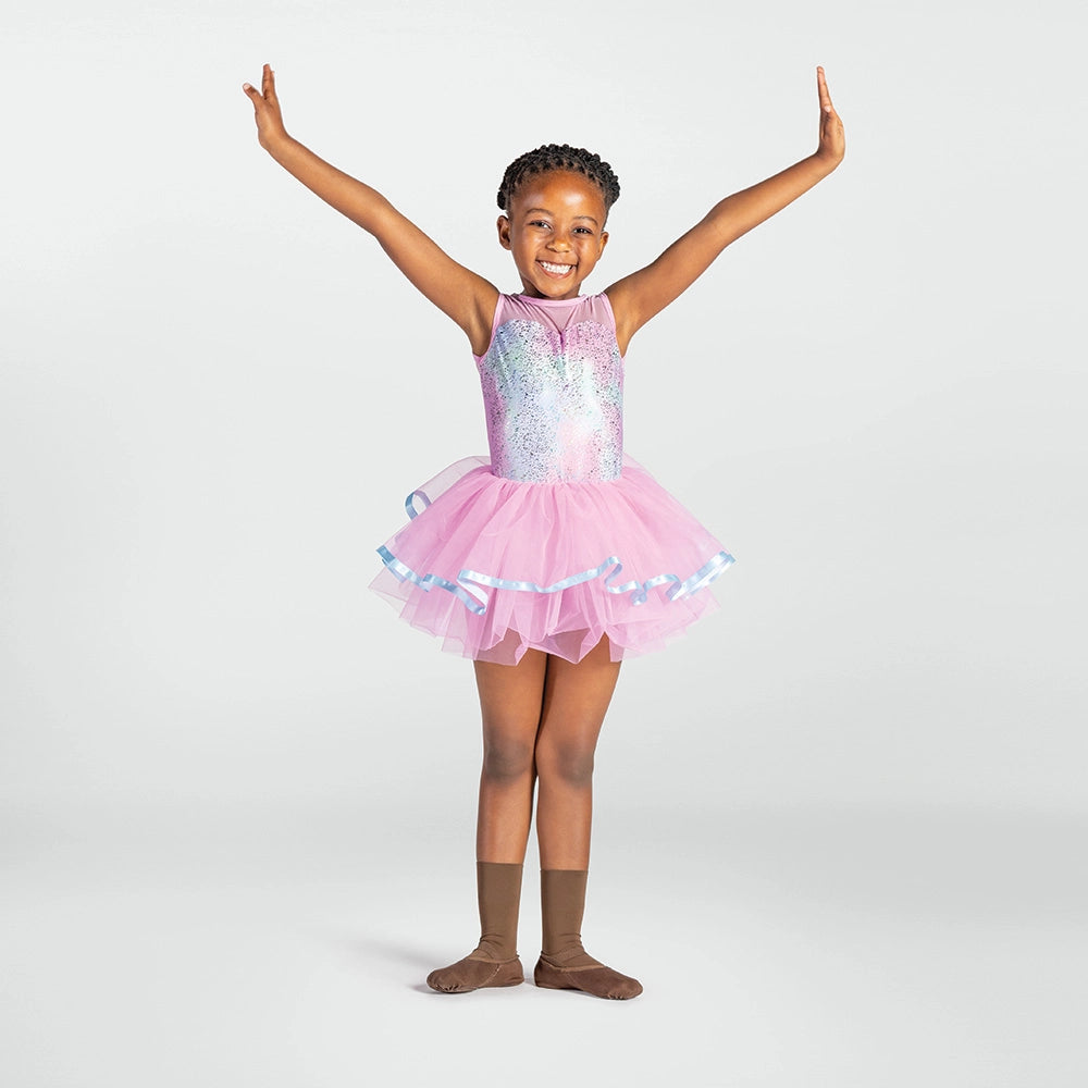 A young girl with braided hair stands in a ballet pose with arms raised, wearing a sparkly lavender and silver sequined leotard with a pink tutu skirt trimmed in light blue, paired with brown dance socks, against a light gray studio background.