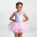 A young girl smiling and wearing a sleeveless ballet leotard with a pink tulle tutu skirt and a glittery iridescent bodice, set against a plain light gray studio background.