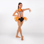 A young girl dancer posing in a sleeveless leotard with an orange and silver glittery corset-style bodice, black shorts, and an orange tulle skirt, set against a plain white studio background.