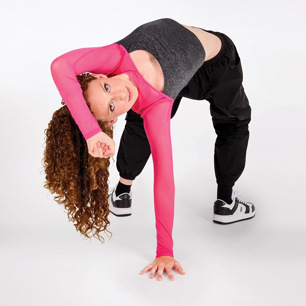 A woman with long curly brown hair poses in a yoga backbend stretch, wearing a bright pink long-sleeve crop top, a dark gray heathered crop top, black sweatpants, and black-and-white sneakers against a solid white studio background.
