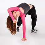 A woman with long curly brown hair poses in a yoga backbend stretch, wearing a bright pink long-sleeve crop top, a dark gray heathered crop top, black sweatpants, and black-and-white sneakers against a solid white studio background.