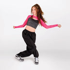 A young woman with curly brown hair poses in a dance or athletic stance, wearing a long-sleeve crop top with a gray ribbed body and bright pink sleeves, paired with loose black sweatpants and black-and-white sneakers, against a plain light gray studio background.
