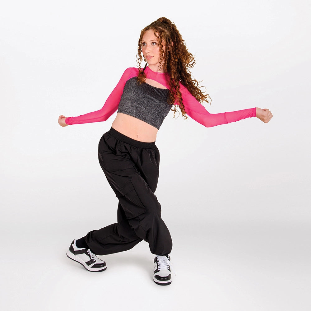 A young woman with curly brown hair poses in a dance or athletic stance, wearing a long-sleeve crop top with a gray ribbed body and bright pink sleeves, paired with loose black sweatpants and black-and-white sneakers, against a plain light gray studio background.