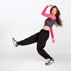 A woman posing in a studio wearing a two-tone long-sleeve crop top with a dark gray heathered body and bright pink sleeves, paired with black sweatpants and black-and-white sneakers.
