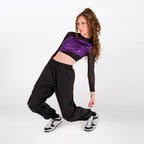 A model with curly brown hair poses in a black and purple dancewear outfit, featuring a long-sleeve crop top with a shiny purple chest panel and black sheer sleeves, paired with loose black pants and black-and-white sneakers, against a plain white studio background.