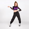 A woman modeling a black long-sleeve crop top with a metallic purple chest panel, paired with black cargo pants and white sneakers, posing against a light gray studio background.