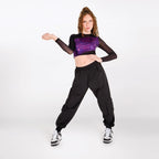 A woman modeling a black long-sleeve crop top with a metallic purple chest panel, paired with black cargo pants and white sneakers, posing against a light gray studio background.