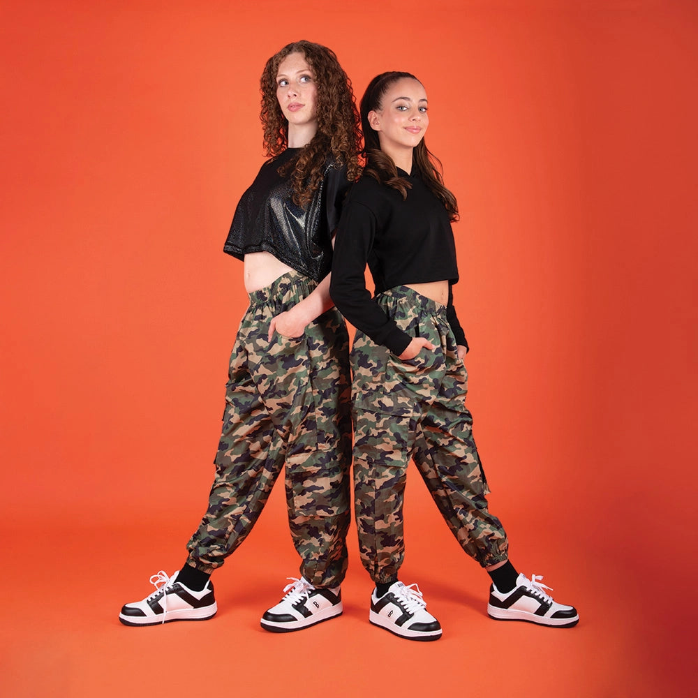 Two young women modeling matching outfits consisting of black cropped long-sleeve shirts and baggy camouflage cargo pants, standing against a solid orange background.
