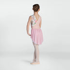A young girl dancer is shown from the back wearing a sleeveless pink leotard with a floral patterned back panel, a pink pleated skirt, and light pink tights against a light gray studio background.