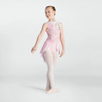 A young girl dancer wearing a light pink sleeveless leotard with a floral print on the back, a matching pink tutu, and light pink tights, posing against a light gray studio background.