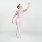 A young girl dancer posing in a light pink sleeveless leotard with floral embroidery, white tights, and ballet slippers against a light gray studio background.