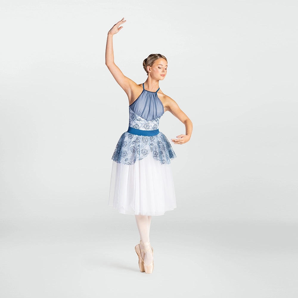 A female ballet dancer posing in a sleeveless blue and white patterned leotard with a white tulle skirt, standing en pointe against a light gray studio background.