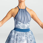 A woman wearing a sleeveless halter dress with a blue-gray ruched bodice, a blue waistband, and a white skirt with a blue floral pattern, shown against a light gray studio background.