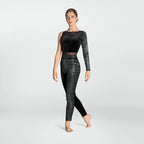 A woman modeling a black two-piece dancewear set consisting of a long-sleeve crop top with sheer mesh shoulders and high-waisted leggings with a textured black-and-white pattern, set against a light gray studio background.