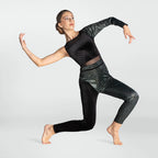 A woman in a black dance leotard and patterned leggings poses dynamically in a lunge position against a light gray studio background.