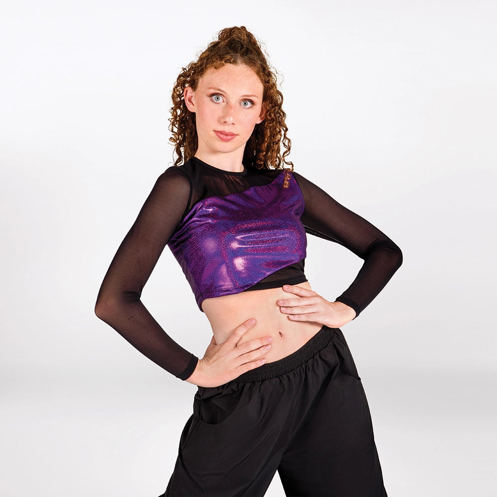 A young woman with curly light brown hair modeling a black and purple dance crop top with sheer long sleeves, paired with black dance pants, against a light gray studio background.