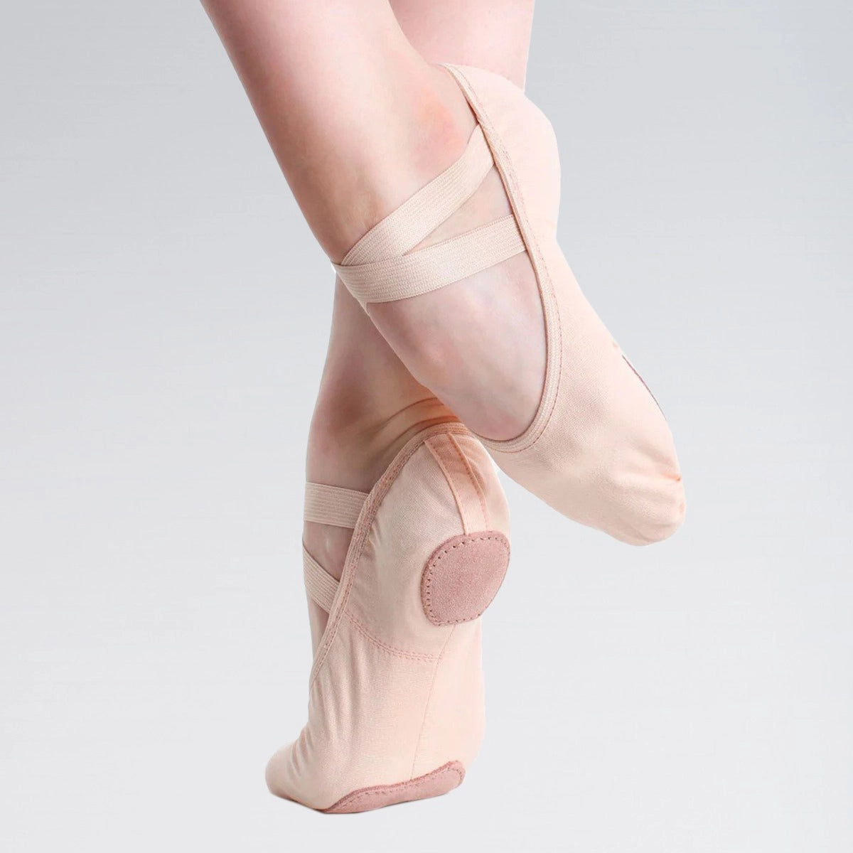 A close-up of a dancer's feet wearing pale blush pink pointe shoes with elastic ankle straps, posed against a light gray studio background.